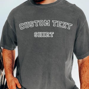 May include: A charcoal gray t-shirt with the words "CUSTOM TEXT" arched above "SHIRT" in white, outlined block letters. The shirt has short sleeves and a relaxed fit.