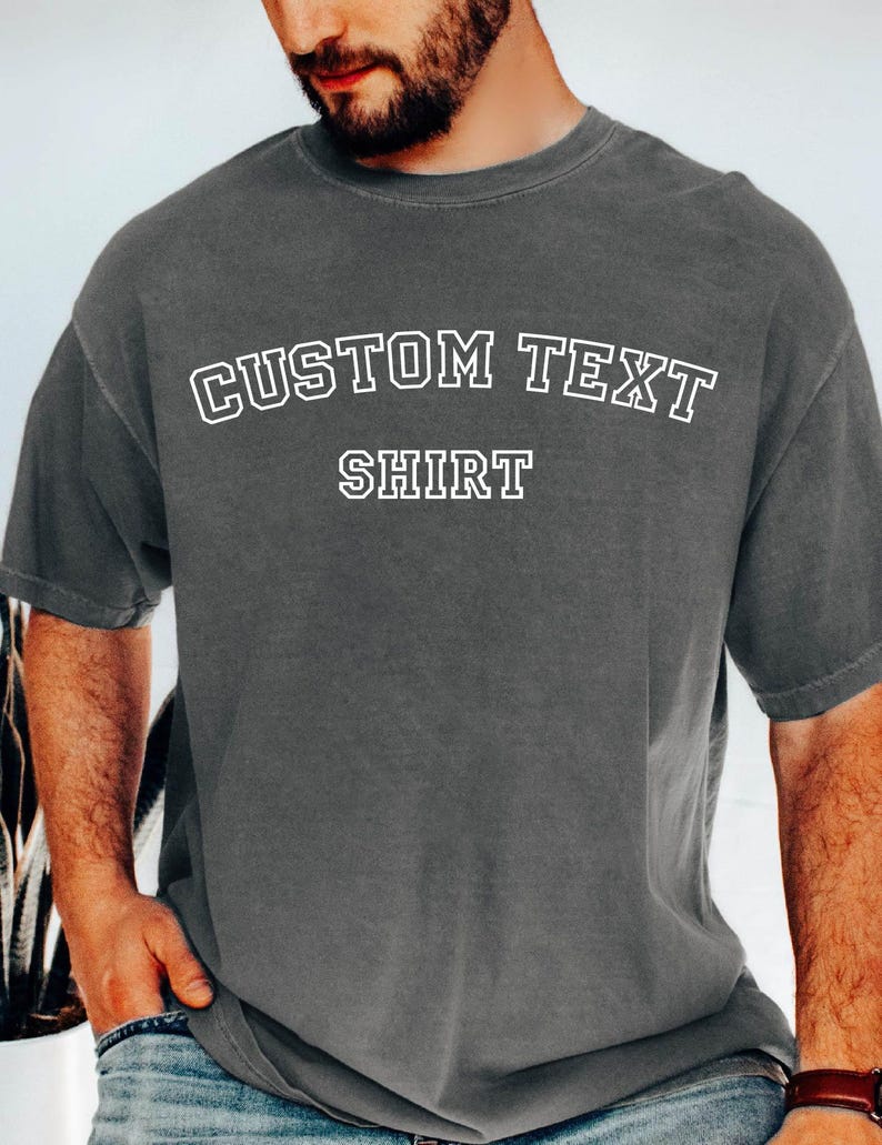 May include: A charcoal gray t-shirt with the words "CUSTOM TEXT" arched above "SHIRT" in white, outlined block letters. The shirt has short sleeves and a relaxed fit.