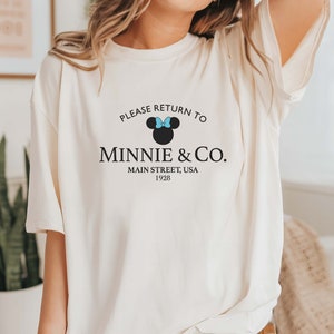 Pu&ograve; includere: Maglietta bianca con una grafica nera che dice "Please return to Minnie & Co. Main Street, USA 1928". La grafica include anche una silhouette nera di Minnie Mouse con un fiocco blu.