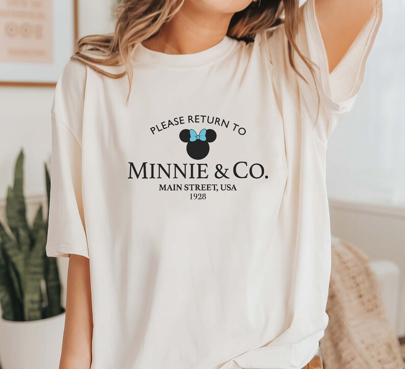 Pu&ograve; includere: Maglietta bianca con una grafica nera che dice "Please return to Minnie & Co. Main Street, USA 1928". La grafica include anche una silhouette nera di Minnie Mouse con un fiocco blu.