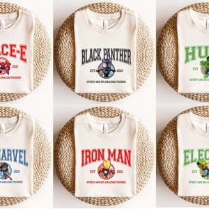 Pu&ograve; includere: Sei t-shirt color crema con diversi design di personaggi di supereroi. Ogni t-shirt presenta una grafica colorata di un supereroe, il nome del supereroe in uno stendardo colorato e il testo "EST 2021 SPIDEY AND HIS AMAZING FRIENDS".