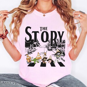 Puede incluir: Camiseta rosa claro con un estampado gr&aacute;fico de personajes de dibujos animados cruzando una calle, con el texto "THE STORY OF TOYS" en negro. El dise&ntilde;o incluye una escena de carretera con coches y conos de tr&aacute;fico.