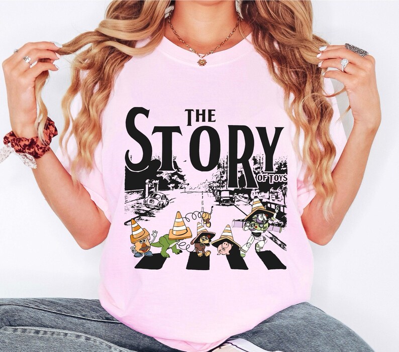 Puede incluir: Camiseta rosa claro con un estampado gr&aacute;fico de personajes de dibujos animados cruzando una calle, con el texto "THE STORY OF TOYS" en negro. El dise&ntilde;o incluye una escena de carretera con coches y conos de tr&aacute;fico.