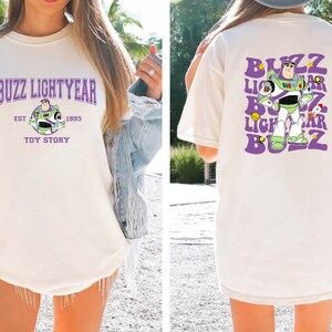 May include: A white t-shirt with a purple and green graphic of Buzz Lightyear from Toy Story. The front of the shirt says "Buzz Lightyear Est. 1995 Toy Story". The back of the shirt has a repeated graphic of Buzz Lightyear with the words "Buzz Lightyear" in a purple, retro font.