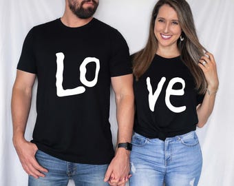 May include: Black t-shirts with the word "Lo" and "ve" in white. The shirts are worn by a couple holding hands. The woman is wearing blue jeans. The man is wearing blue jeans and a black watch.