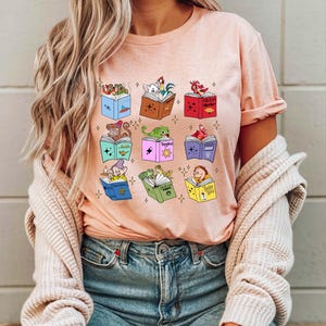 May include: A pink t-shirt with a graphic print of various Disney characters reading books. The books have titles like "Mulan", "Aladdin", "Peter Pan", and "Beauty and the Beast".