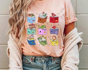 Op de afbeelding: Een roze T-shirt met een grafische print van verschillende Disney-figuren die boeken lezen. De boeken hebben titels zoals "Mulan", "Aladdin", "Peter Pan" en "Belle en het Beest".