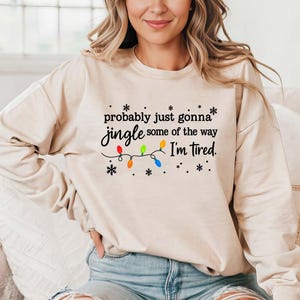 May include: A beige sweatshirt with the text "probably just gonna jingle some of the way I'm tired." The text is in black, with colorful Christmas lights and snowflakes. The sweatshirt is long-sleeved.