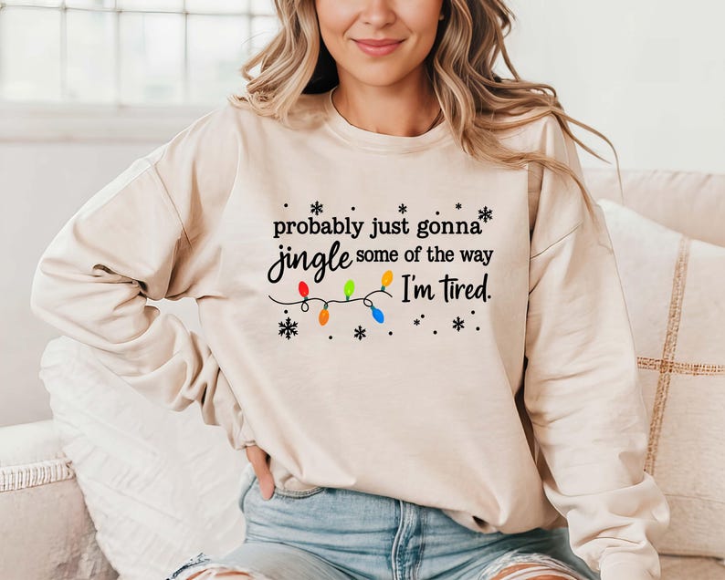 May include: A beige sweatshirt with the text "probably just gonna jingle some of the way I'm tired." The text is in black, with colorful Christmas lights and snowflakes. The sweatshirt is long-sleeved.