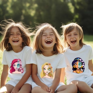 May include: Three children wearing white t-shirts with cartoon images of Disney princesses with sugar skull makeup. The girls are smiling and looking at the camera.