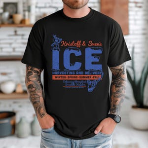 May include: Black t-shirt featuring the text "Kristoff & Sven's ICE" in blue and orange. The design includes images of Olaf and Sven, with text indicating ice harvesting and delivery throughout Arendelle Kingdom. The shirt is a crew neck.