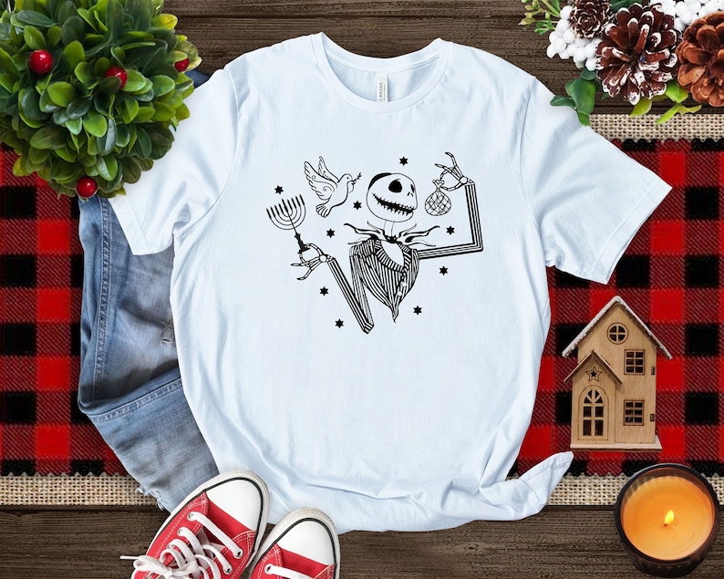 May include: A light blue t-shirt featuring a black and white graphic of a skeleton holding a menorah and a Christmas bauble. The skeleton is wearing a striped shirt and a bow tie. There are stars around the skeleton.
