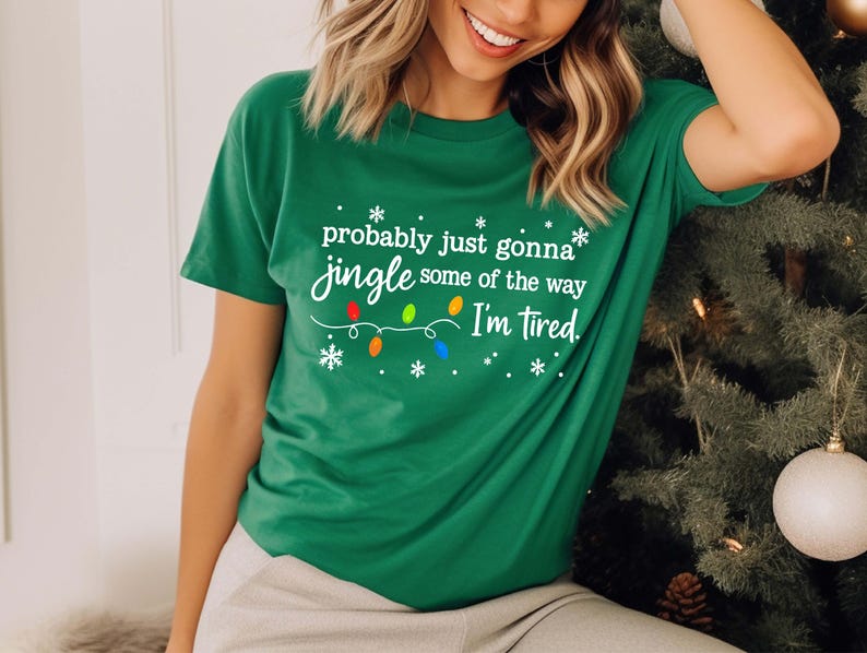 May include: A green t-shirt with the text "probably just gonna jingle some of the way I'm tired." The text is in white with colorful Christmas light accents. The shirt is a festive holiday apparel item.