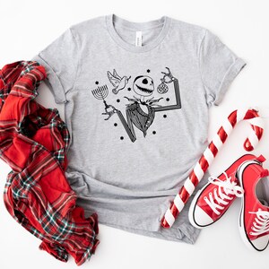 May include: A grey t-shirt featuring a black and white illustration of Jack Skellington from The Nightmare Before Christmas holding a menorah and a candy cane. The shirt is surrounded by red and white candy canes, a red checked shirt, and red trainers.