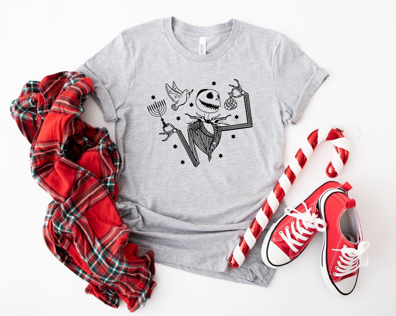 May include: A grey t-shirt featuring a black and white illustration of Jack Skellington from The Nightmare Before Christmas holding a menorah and a candy cane. The shirt is surrounded by red and white candy canes, a red checked shirt, and red trainers.