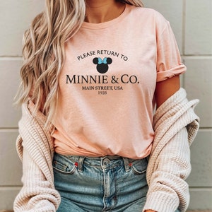 Pu&ograve; includere: Una maglietta rosa con una grafica nera di Minnie Mouse con un fiocco blu. La grafica dice "Please Return To Minnie & Co. Main Street, USA 1928".