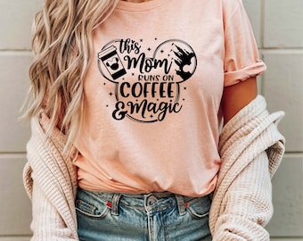 May include: A light peach-colored t-shirt with the text "This Mom Runs on Coffee & Magic" in black. The design includes a coffee cup, Mickey Mouse ears, and a castle silhouette. The shirt is made of soft material.