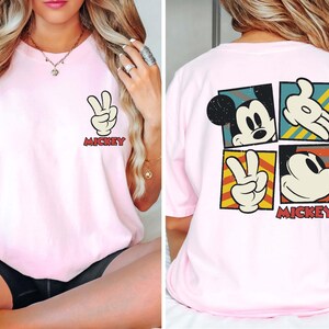 Pode incluir: Camiseta rosa com um gr&aacute;fico do Mickey Mouse na frente e um gr&aacute;fico do Mickey Mouse maior nas costas. O gr&aacute;fico nas costas apresenta o rosto do Mickey Mouse, uma m&atilde;o fazendo um sinal de paz e outra m&atilde;o fazendo um sinal de paz. O texto "Mickey" est&aacute; impresso abaixo do gr&aacute;fico.