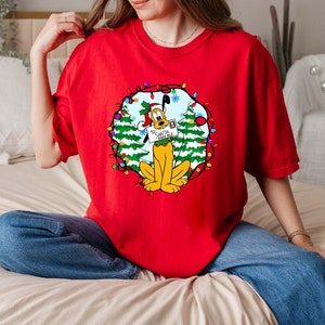 May include: Red t-shirt featuring a Christmas-themed graphic of Pluto wearing a Santa hat and holding a letter addressed to Santa. The graphic is surrounded by a wreath of green trees and colourful lights.