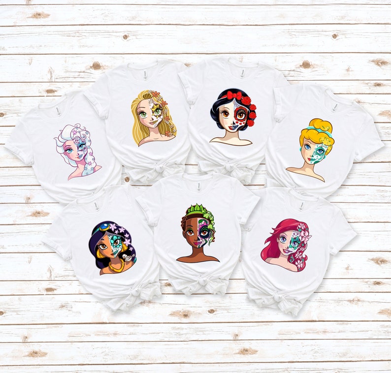 May include: Six white t-shirts with different Disney princess characters on them. Each princess has a sugar skull design on half of their face. The princesses are: Elsa, Rapunzel, Snow White, Cinderella, Jasmine, and Ariel.