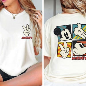 Pode incluir: Camiseta branca com um gr&aacute;fico do Mickey Mouse na frente e nas costas. O gr&aacute;fico da frente &eacute; uma &uacute;nica m&atilde;o do Mickey Mouse fazendo um sinal de paz com o texto "Mickey" abaixo. O gr&aacute;fico das costas &eacute; uma colagem de quatro imagens do Mickey Mouse, incluindo um sinal de paz, uma cabe&ccedil;a do Mickey Mouse e uma m&atilde;o do Mickey Mouse fazendo um sinal de paz. O texto "Mickey" est&aacute; abaixo da colagem.