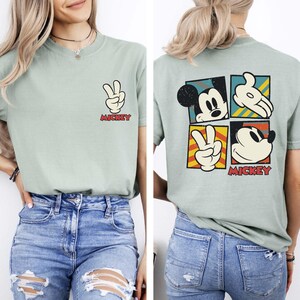 Pode incluir: Uma camiseta verde claro com um gr&aacute;fico vintage do Mickey Mouse nas costas. O gr&aacute;fico apresenta o Mickey Mouse em quatro poses diferentes, incluindo um sinal de paz. A camiseta tamb&eacute;m tem a palavra "Mickey" impressa na frente e atr&aacute;s.
