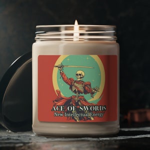Op de afbeelding: Een witte kaars met een rood, groen en geel etiket waarop een skelet met een zwaard te zien is. Het etikett  leest "Ace of Swords New Intellectual Energy".