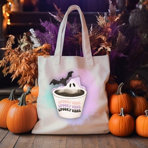 Puede incluir: Bolsa de lona blanca con un diseño de fantasma y murciélago. La bolsa tiene el texto "Spooky Vibes" repetido tres veces. La bolsa está rodeada de calabazas naranjas.