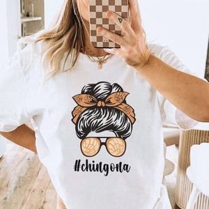 May include: White t-shirt featuring a graphic of a woman's hair styled in a bun, sunglasses, and a headband. The text "#chingona" is below the graphic. The person in the image is wearing a brown beanie and holding a phone.