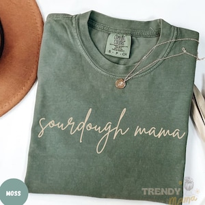 May include: A moss green t-shirt with the words "sourdough mama" in a cursive font. A silver necklace with a small pendant rests on the shirt. The shirt is folded and has a Comfort Colors tag.