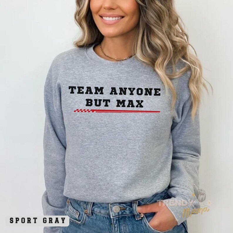 Peut inclure: Un sweat-shirt gris avec le texte "TEAM ANYONE BUT MAX" en lettres noires. Une ligne rouge avec un motif &agrave; carreaux se trouve sous le texte. Le sweat-shirt est port&eacute; par une personne aux cheveux longs et bruns.