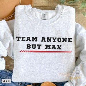 Peut inclure: Un sweat-shirt gris avec le texte "TEAM ANYONE BUT MAX" en lettres noires avec une ligne &agrave; carreaux rouge et blanche en dessous. Le sweat-shirt est pos&eacute; sur une surface en bois avec un chapeau marron et un jean bleu.