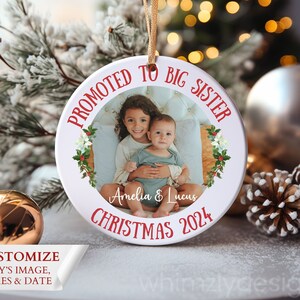 May include: A white ceramic Christmas ornament with a photo of two children. The ornament reads "Promoted to Big Sister" in red, with the names "Amelia & Lucus" and "Christmas 2024". A gold ribbon is attached for hanging. The ornament is surrounded by festive decorations.