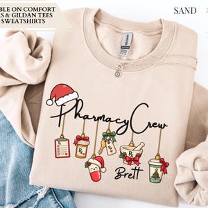 May include: A sand-colored crewneck sweatshirt featuring a graphic print with the text Pharmacy Crew and Brett. The design includes hanging ornaments shaped like a prescription pad, pill bottle, keys, mortar and pestle, and a coffee cup.