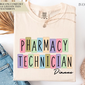 May include: An ivory t-shirt featuring the text PHARMACY TECHNICIAN in black capital letters, with each letter enclosed in a pastel-colored rectangular box. The name Dianne is printed in black script below the text. The shirt has a crew neckline.
