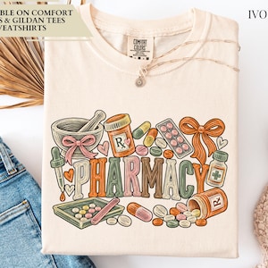 May include: An ivory t-shirt featuring a colorful graphic print with the word PHARMACY in large block letters. The design includes illustrations of a mortar and pestle, pill bottles, various capsules, tablets, a dropper, and a bow.