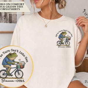 May include: An ivory crewneck t-shirt featuring a graphic of a green frog riding a blue bicycle. The text reads "Off to Turn Can't into Can" and "Brianna - OTR/L". The frog wears a blue shirt and tan pants, with a basket containing tools on the bike.