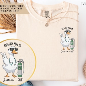 May include: Ivory-colored t-shirt with a cartoon goose wearing sunglasses and a stethoscope, holding an oxygen tank. The text "Airway Ninja" and "Jessica - RRT" are printed on the shirt. The shirt is available on Comfort Colors & Gildan tees and sweatshirts.