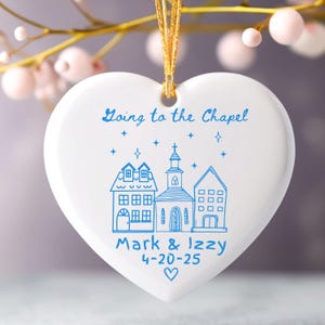 May include: White heart-shaped ornament with blue text and illustrations. The ornament says "Going to the Chapel" and features drawings of buildings, stars, and the names "Mark & Izzy" with a date and heart symbol. A gold ribbon is attached for hanging.