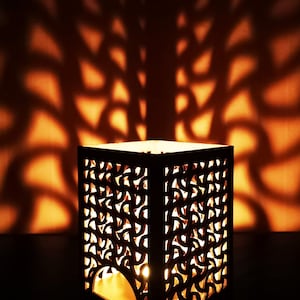 May include: A wooden candle lantern with a intricate cut-out design casts a warm glow, creating a beautiful shadow pattern on the wall behind it.