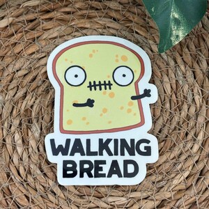 Puede incluir: Una pegatina con una imagen de dibujos animados de una rebanada de pan con una cara de zombi. El pan es amarillo con una corteza roja, ojos negros y una boca cosida. Las palabras "WALKING BREAD" están impresas en negrita, en letras negras debajo de la imagen del pan.