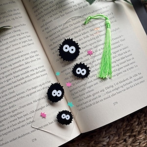 May include: An open book with black, round, cartoon-like bookmarks with white eyes. A green tassel bookmark and pink and blue star-shaped confetti are also present.