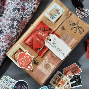 Pode incluir: Um pacote de papel castanho embrulhado com barbante e decorado com vários adesivos. Os adesivos incluem um design floral, um saquinho de chá, um adesivo com tema de livros com o texto "Drink Tea Read Books" e uma etiqueta que diz "Roomance".
