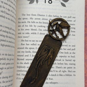 May include: A bronze bookmark featuring a stylized image of a mockingjay, a symbol from the Hunger Games book series. The bookmark is placed on a page of a book with the number 18 visible. The text on the page reads "The boy from District 1 dies before he can pull out the spear. My arrow drives deep into the center of his neck. He falls to his knees and the remainder of his life by yanking of his own blood. I'm reloading side to side, while I shout at the sky. Are there more?"