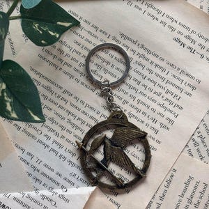 Könnte beinhalten: Ein silberner Schlüsselanhänger mit einem bronzenen Mockingjay-Anhänger. Der Mockingjay ist ein Symbol aus der Buchreihe Die Tribute von Panem. Der Schlüsselanhänger liegt auf einem Hintergrund aus Buchseiten.