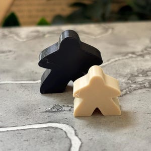 May include: Two board game meeples, one larger black meeple and one smaller beige meeple, standing upright. The figures have a classic human-like shape with rounded heads, outstretched arms, and two legs, positioned together on a flat surface.