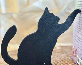 May include: A black metal cat-shaped bookend. The cat is in a playful pose, reaching up with one paw to hold a book in place. The bookend is on a woven surface.