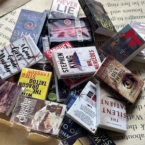 Puede incluir: Una colección de libros en miniatura con varios títulos y autores, incluyendo Stephen King y Agatha Christie. Los libros presentan portadas coloridas y texto, creando un tema literario. Los libros están dispersos sobre un fondo de papel envejecido.