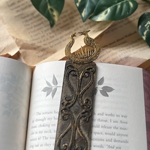 May include: A gold-colored metal bookmark featuring two stylized peacocks facing each other. The bookmark has a detailed design with intricate patterns and a textured surface.