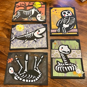 May include: Five Halloween greeting cards with black skeletons of different animals on colorful backgrounds. The animals include a rhinoceros, an owl, a shark, a sloth, and a snake.